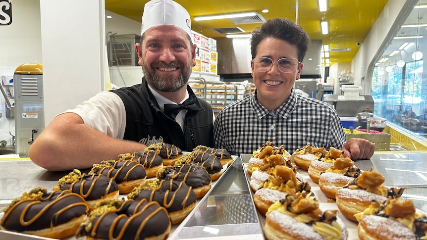 two people standing in front of a tray of delicious looking doughnuts