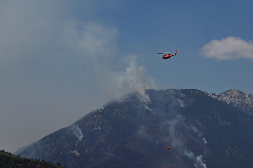 A bright red helicopter is seen from a distance flying over a smoky hillside, hauling a payload at the end of a long cable.