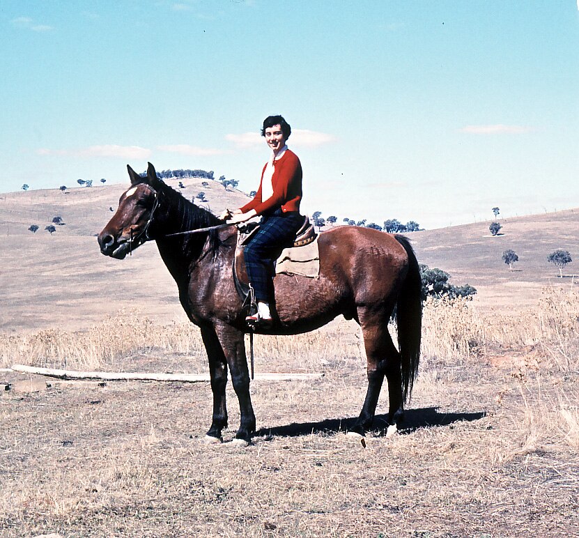 A film photo of a teenage girl sitting on a brown horse, with rolling brown hills and trees in the background.