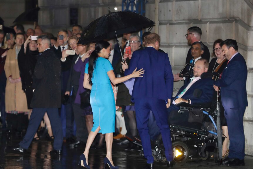 Britain's Prince Harry and Meghan speak to a man in a wheelchair in a crowd of people standing outside in the rain.