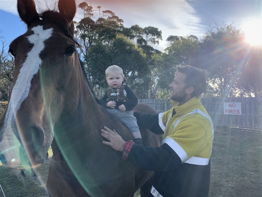 A toddler sitting on the back of a horse while being supported by a man, who is smiling.