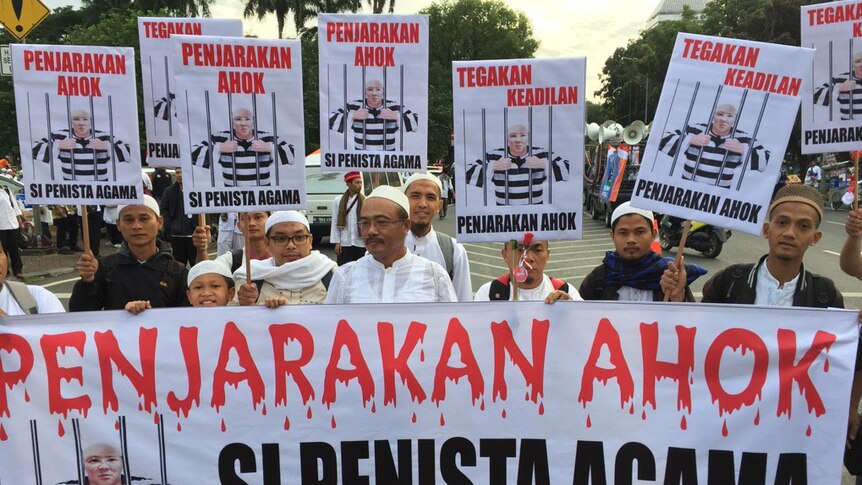 A group of protesters hold one large banner, while holding one smaller one each, that reads "Jail Ahok" in red writing.