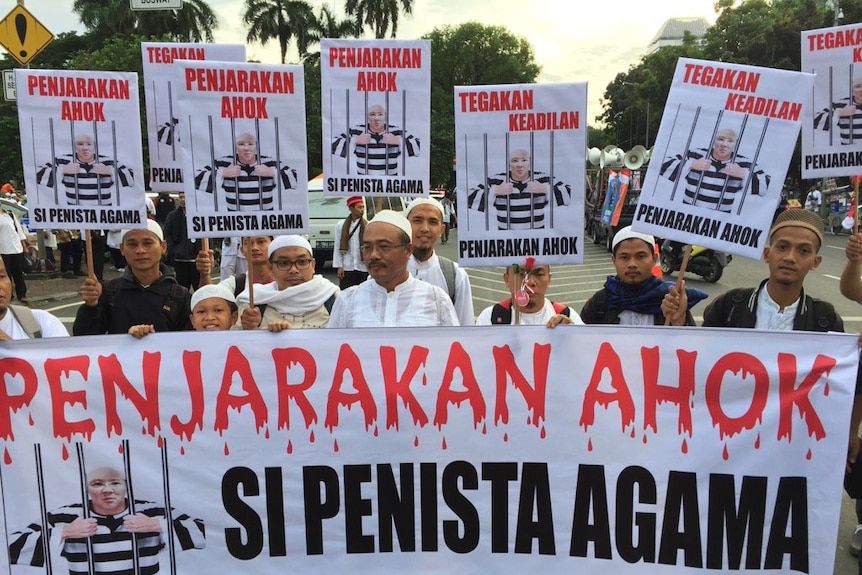 A group of protesters hold one large banner, while holding one smaller one each, that reads "Jail Ahok" in red writing.