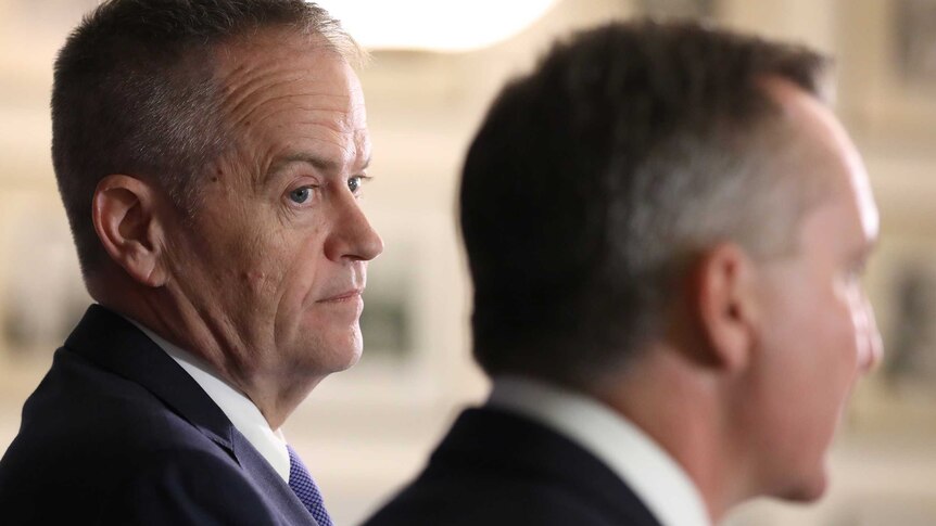 Bill Shorten looks at Chris Bowen, who is only seen from behind. Mr Shorten is wearing a navy suit and purple tie.