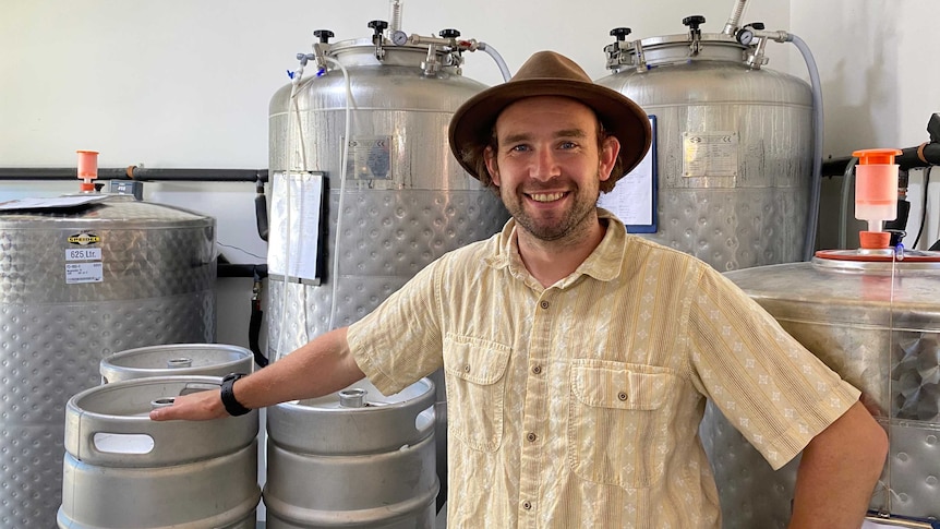 a man wearing a yellow shirt and a brown hat stands in front of some kegs and vats