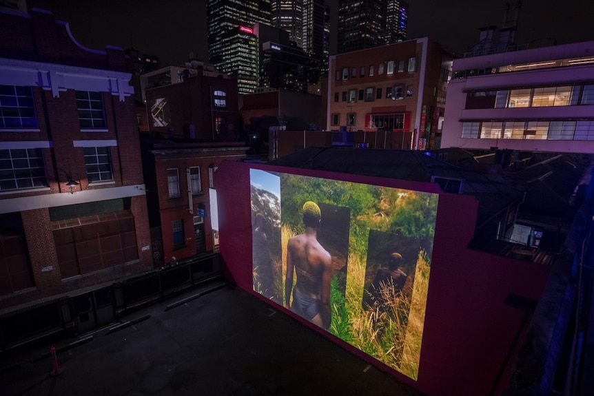 At a rooftop car park, a video is projected onto the wall of a building at night. In the video an African man stands in a field