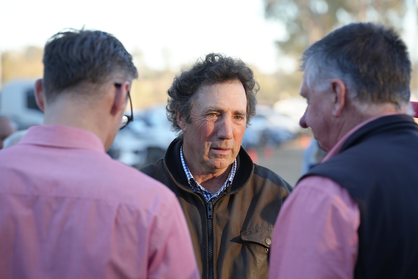 A middle-aged man with dark, curly hair stands outdoors and speaks to two men.