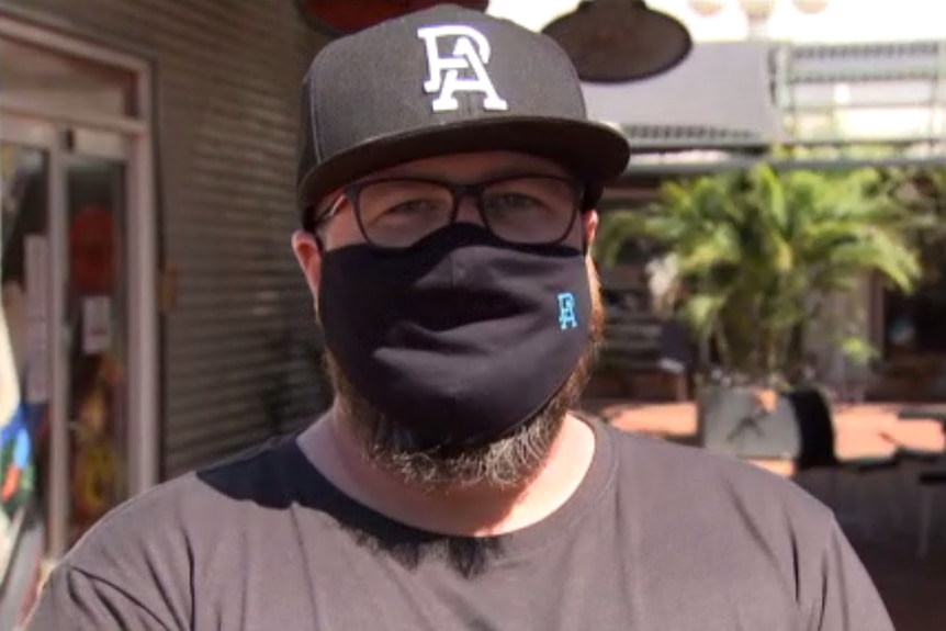 A man with a beard and glasses, wearing a black cap, t-shirt and face mask.