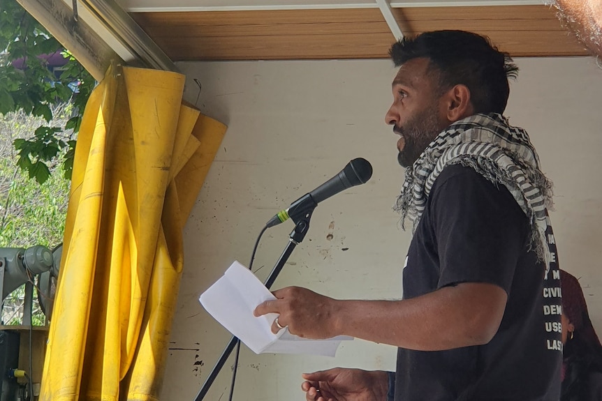 Comedian Nazeem Hussain speaks on stage at a rally, wearing a Palestinian keffiyeh around his neck.