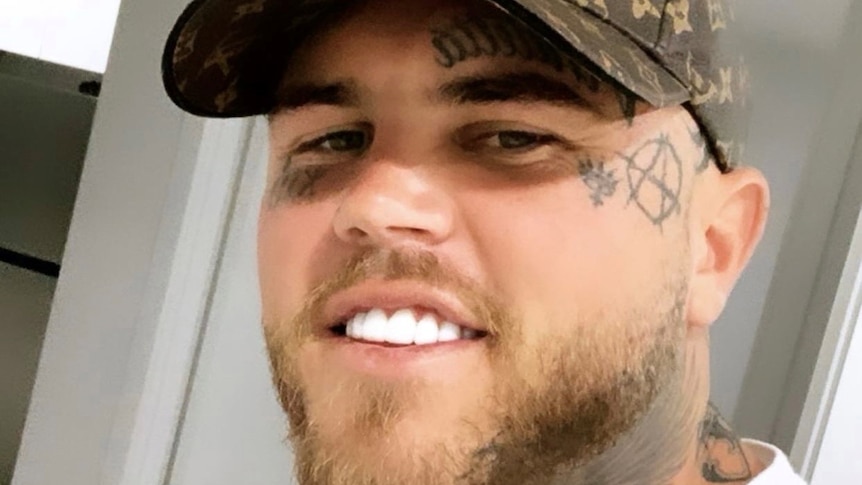 A man with white teeth and a cap as well as facial tattoos smiles at a camera