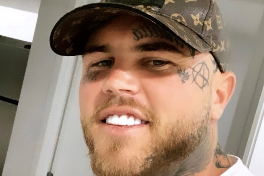 A man with white teeth and a cap as well as facial tattoos smiles at a camera