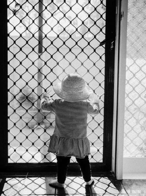 Black and white photo of child in hat looking out a screened door