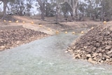 Shallow water flows over rocks on a 5 metre-wide creekbed. Big piles of rocks are on either side of the creek.