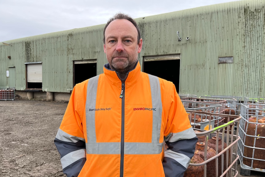A man frowns at the camera while wearing an orange vest in front of a green warehouse.