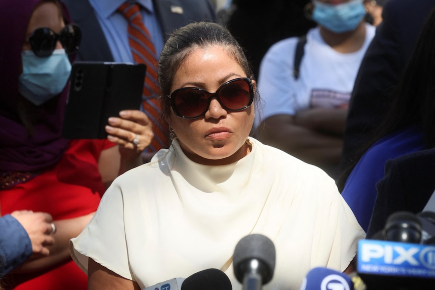 A woman wearing sunglasses speaks to a group of media. 