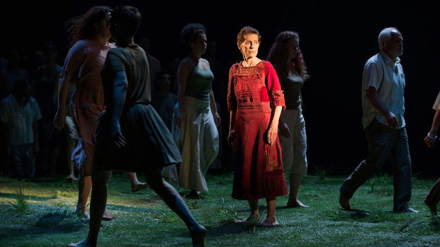 Actress Helen Morse wearing red dress and with short-cropped hair, on stage surrounded by performers.