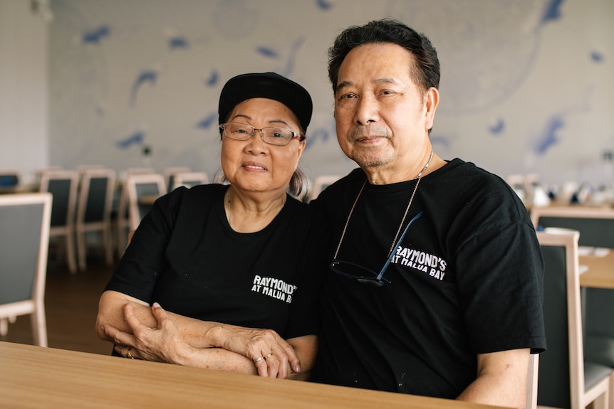 A middle-aged Asian woman and man sit at a table in an empty restaurant as they pose for a photograph.