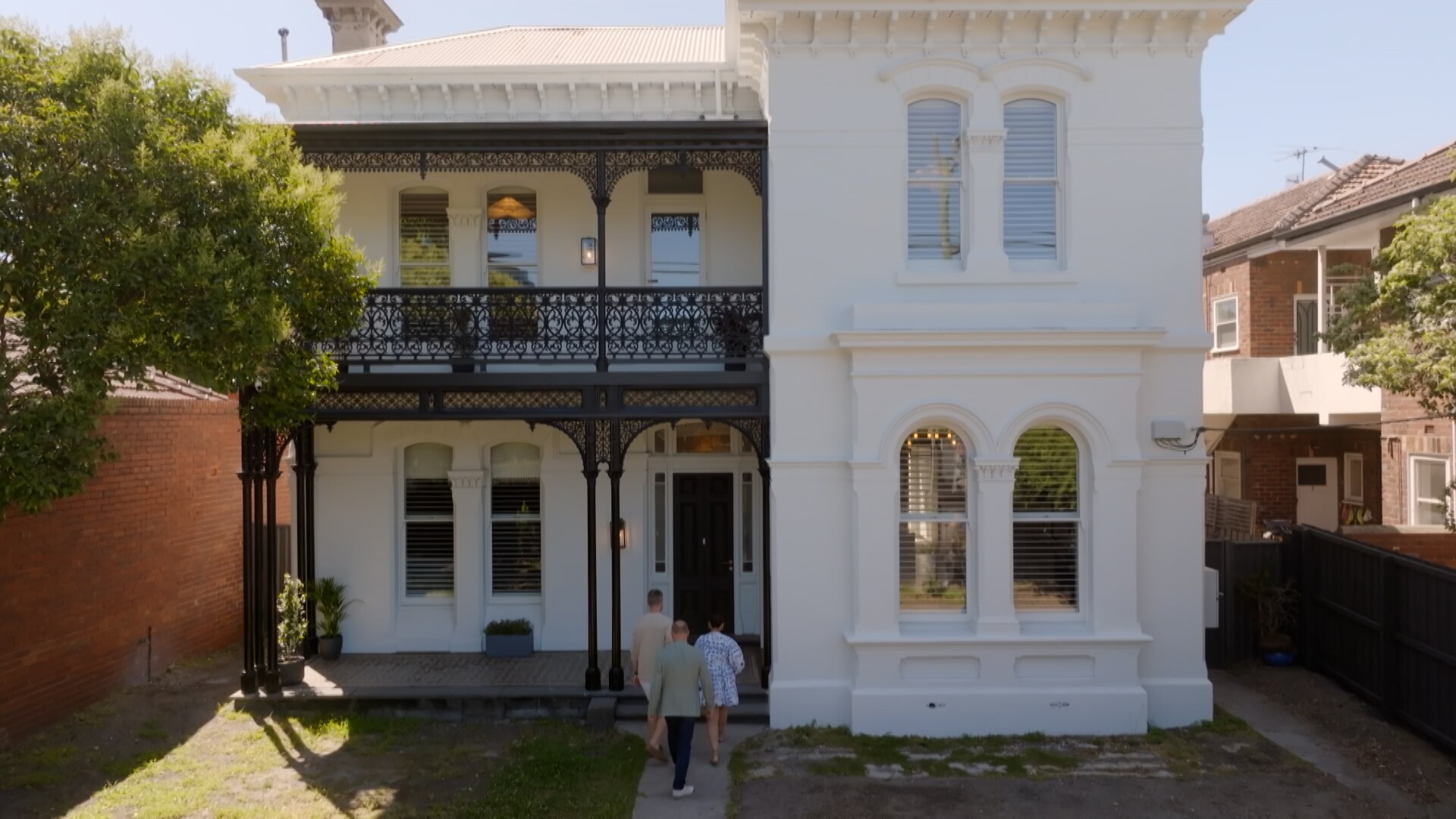 Самостоятельно поддерживающий белый викторианский дом с черными коваными железными балюстрадами с левой стороны. Люди входят