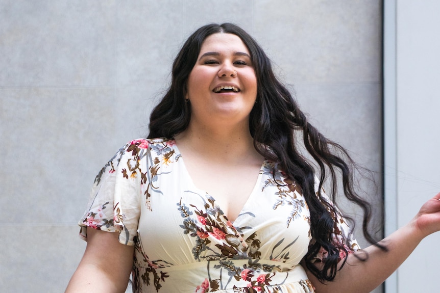 A young woman wearing a floral dress stands in front of a concrete wall, smiling