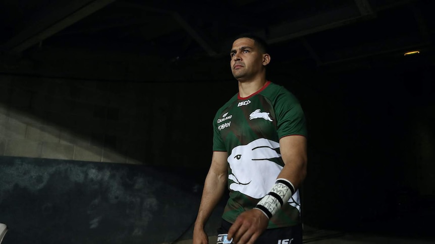 NRL player Cody Walker walks out of a dark change room tunnel wearing his green and red Rabbitohs uniform, looking sombre