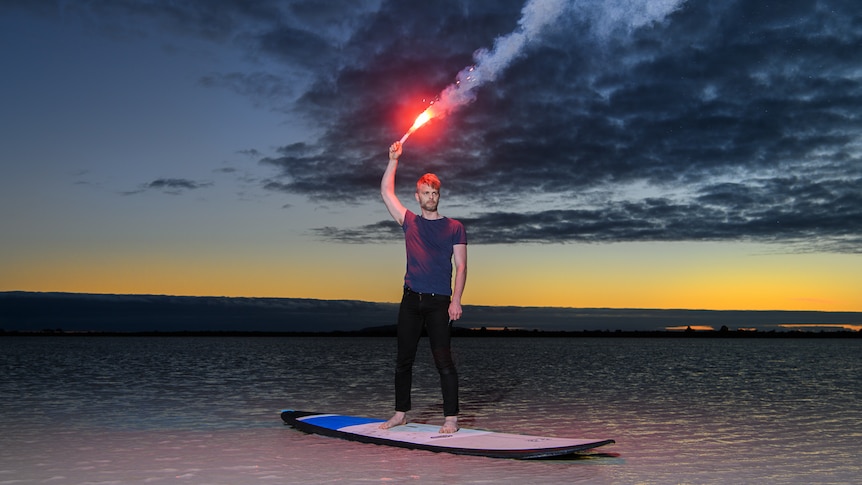 David Finnigan standing on a surfboard holding a flare above his head on a beach at sunset.