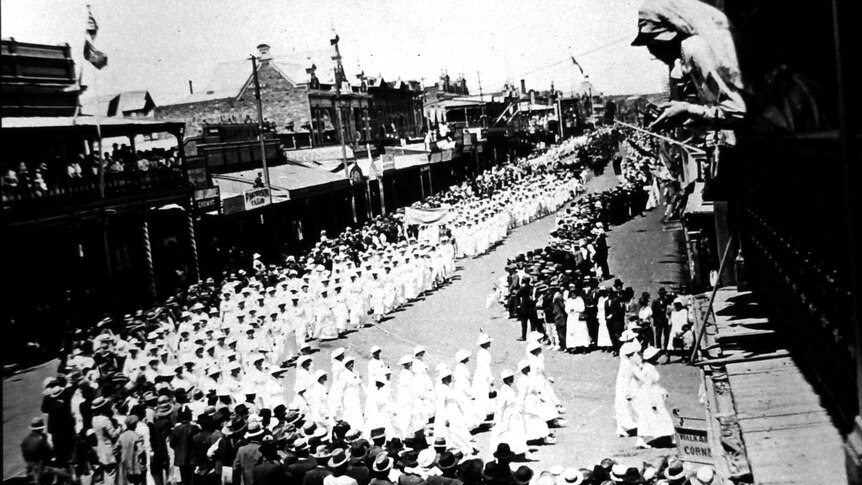A black and white photograph of hundreds of women dressed in white marching a street lined with people.
