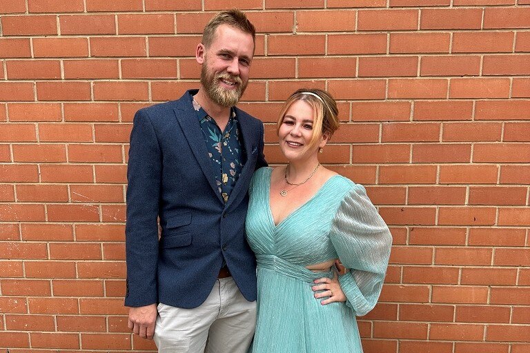 Claire, in a blue dress, and Josh, in a blazer, stand together embracing and posing for a photo
