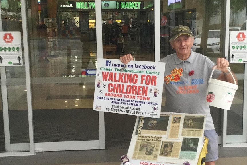 An older man in a cap stands at the entrance to a mall, holding up a sign calling for donations.