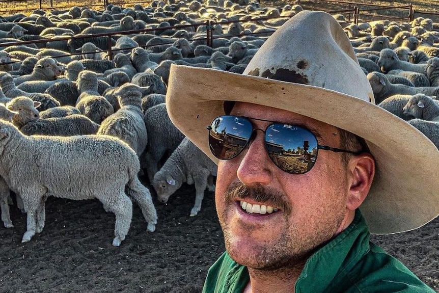 Many with sunglasses and hat takes selfie in front of sheep