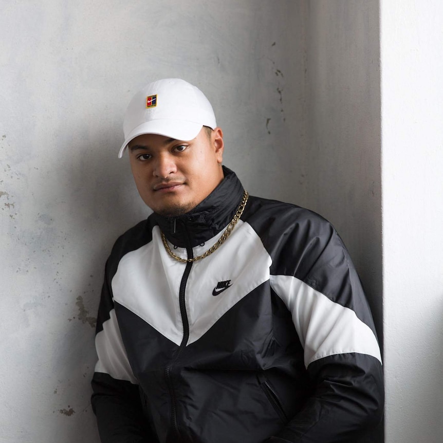 Spenny, a drill rapper, standing against a concrete wall wearing a Nike tracksuit and cap with a gold chain