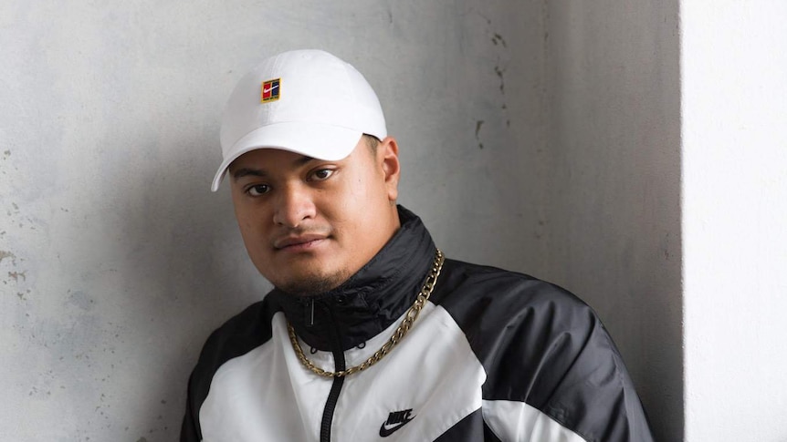 Spenny, a drill rapper, standing against a concrete wall wearing a Nike tracksuit and cap with a gold chain