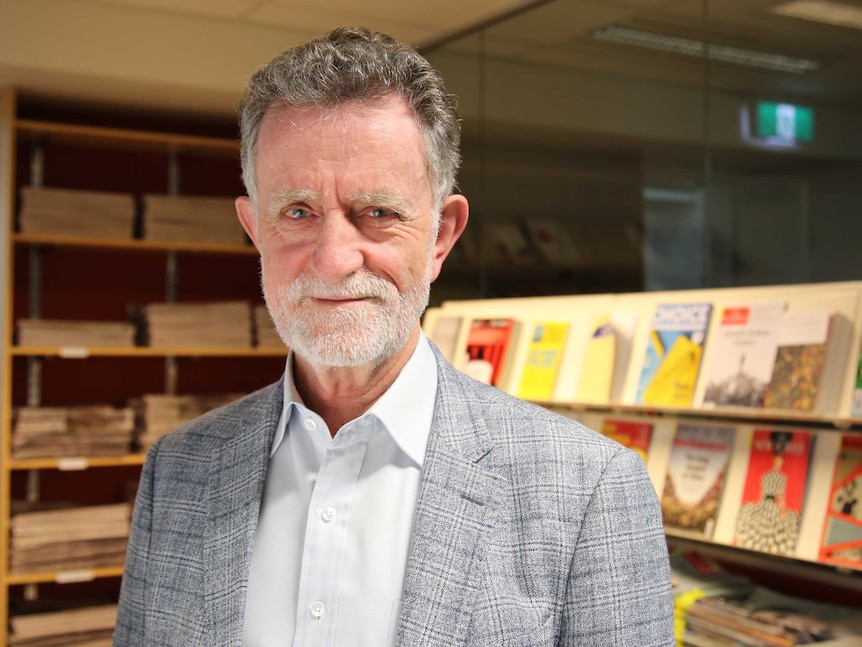 A smiling old man in a gray suit, white shirt, gray beard stands in front of a glass wall with book shelves. 