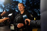 A white, female police officer in uniform stands and speaks with reporters holding phones and microphones.
