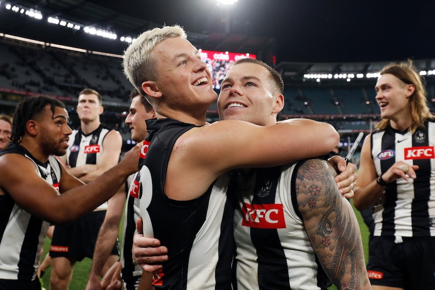 Jack Ginnivan and Jamie Elliott hug while surrounded by Collingwood teammates after an AFL win.