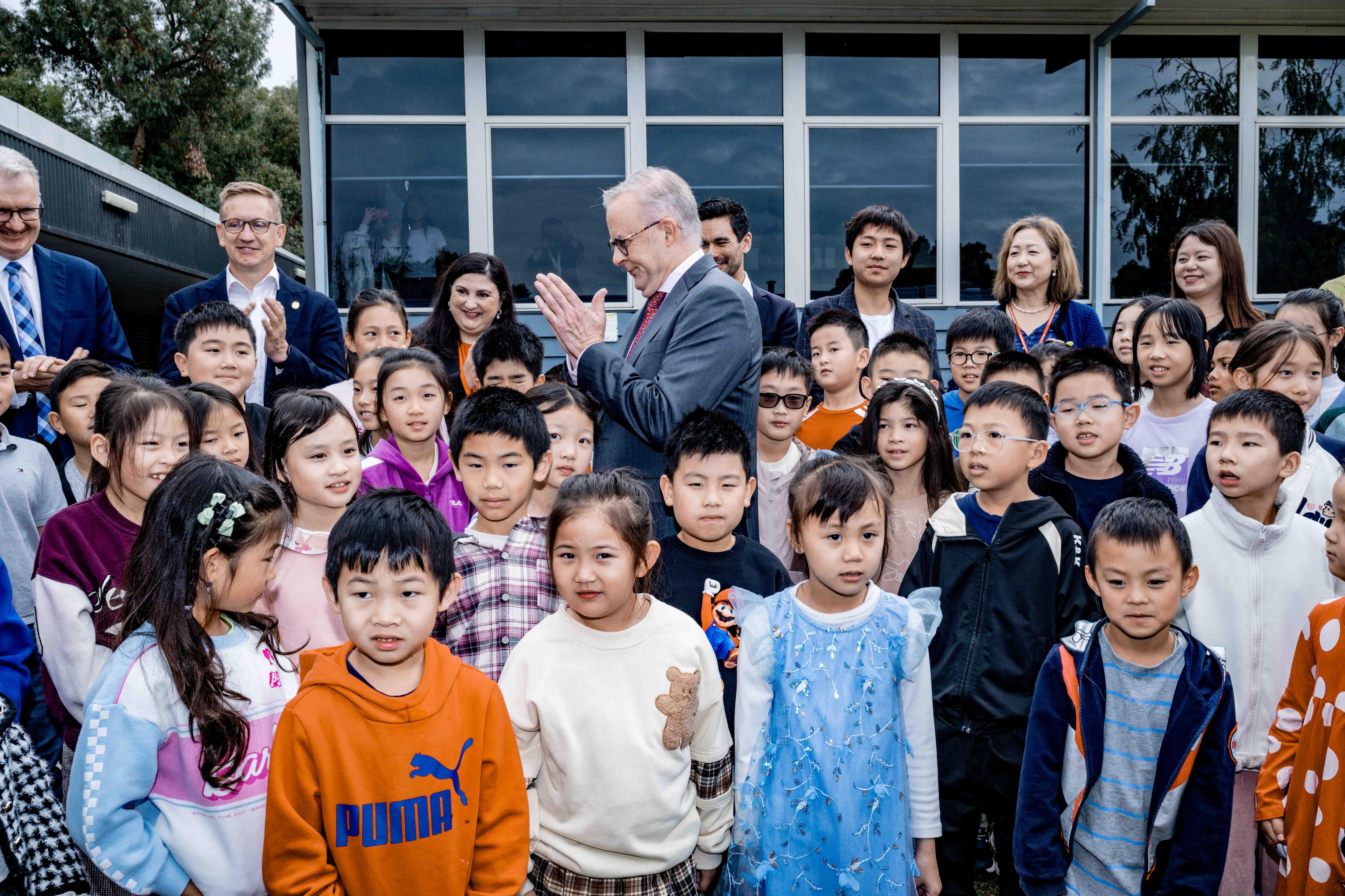Albanese stands in the middle of a group of children, he&#x27;s looking to the side with palms pressed together in greeting