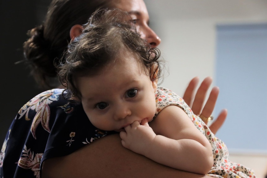 A baby with brown eyes looks at the camera over mum's shoulder.