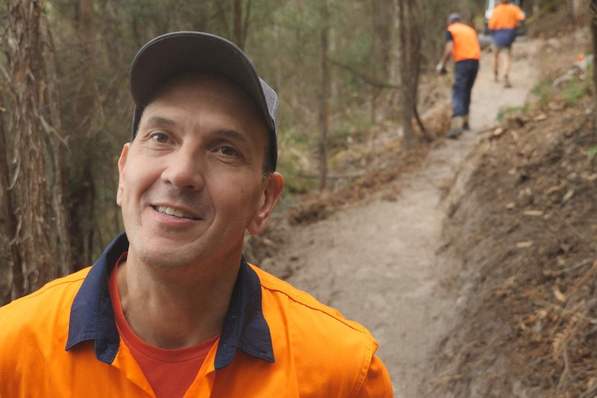 Man smiling close to camera in hi vis vest, workers building mtb trail behind