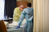 Two men in personal protective equipment stand by a patient's bed.