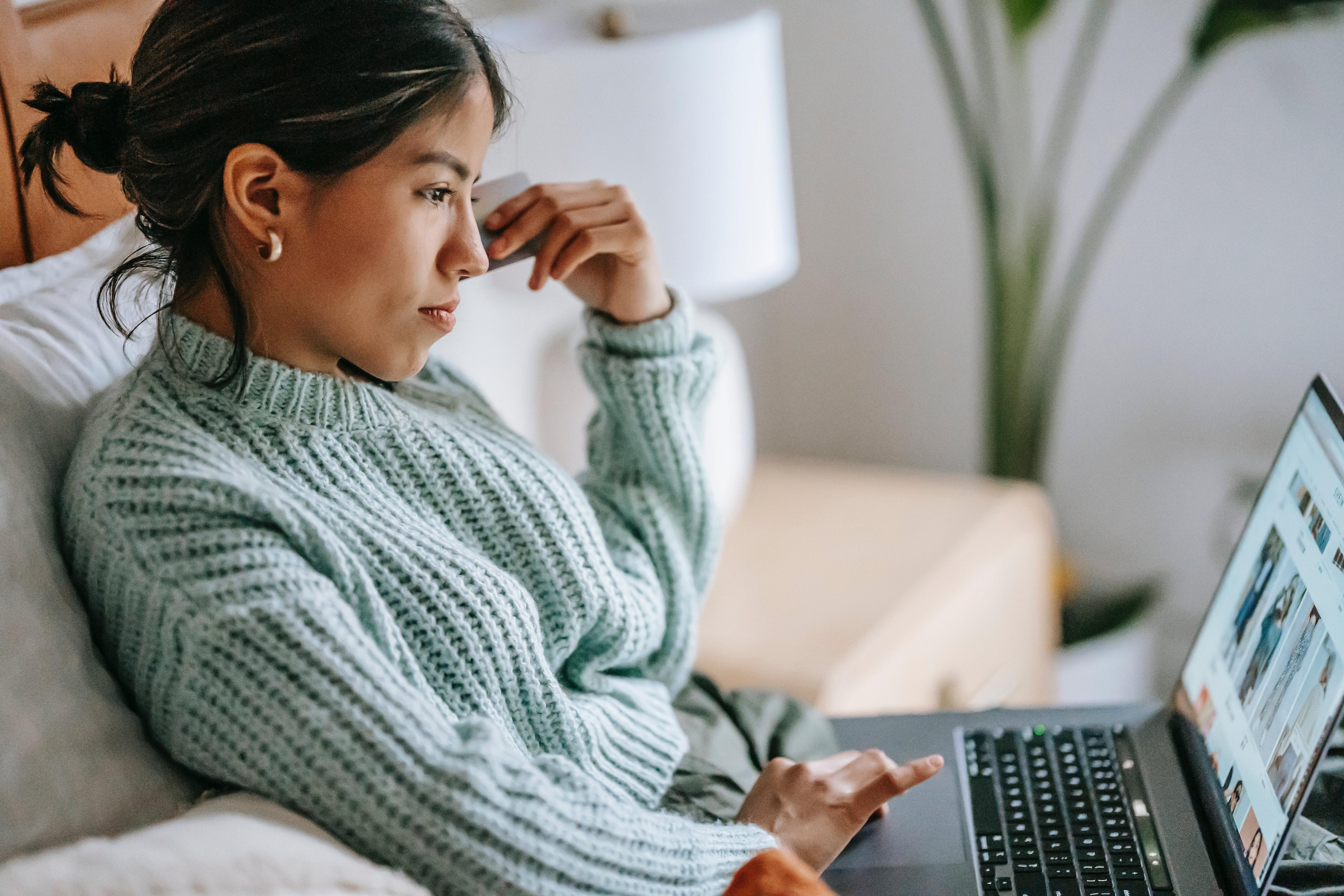 Woman in teal jumper looking at her computer on the couch, a credit card in hand.