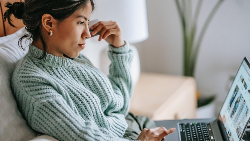 Woman in teal jumper looking at her computer on the couch, a credit card in hand.