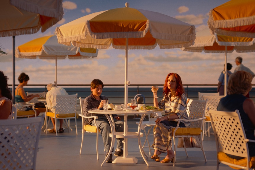 A film still of Armen Nahapetian, a teen boy, and Zoe Lister-Jones, a red-haired woman, sitting under an umbrella on a ship deck