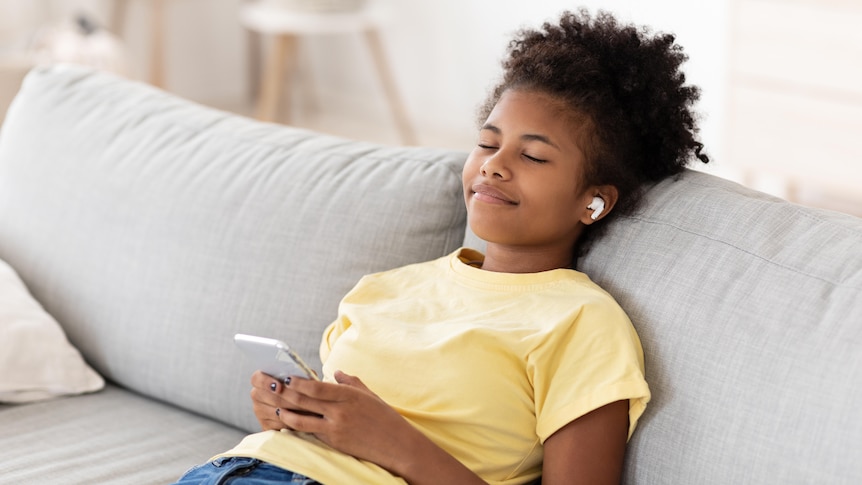 A young black girl sits back on a sofa and smiles while listening to music through earbuds.