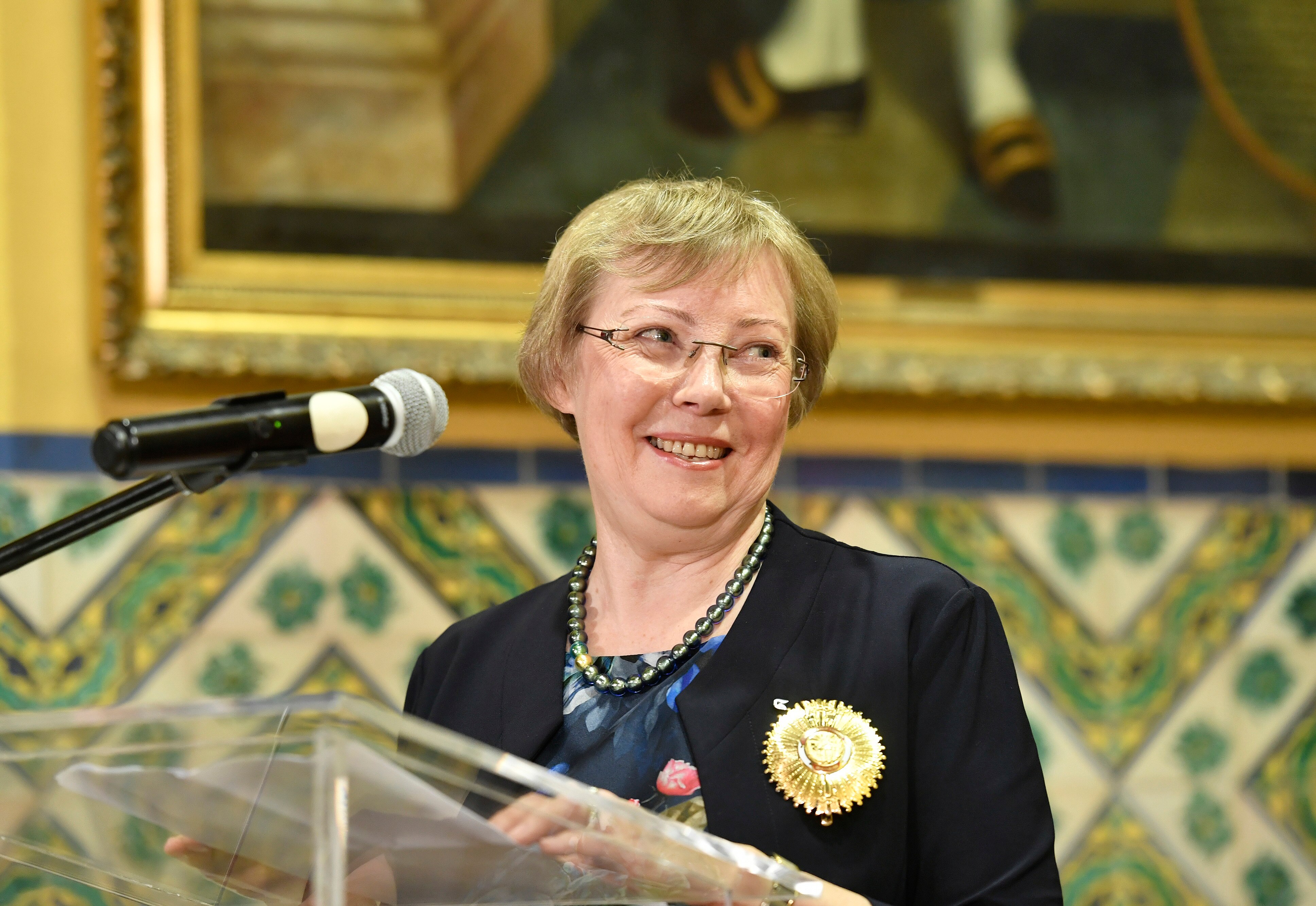 A woman with cropped blonde hair and glasses smiles at a lectern 