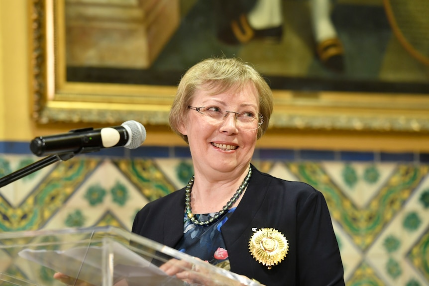 A woman with cropped blonde hair and glasses smiles at a lectern 