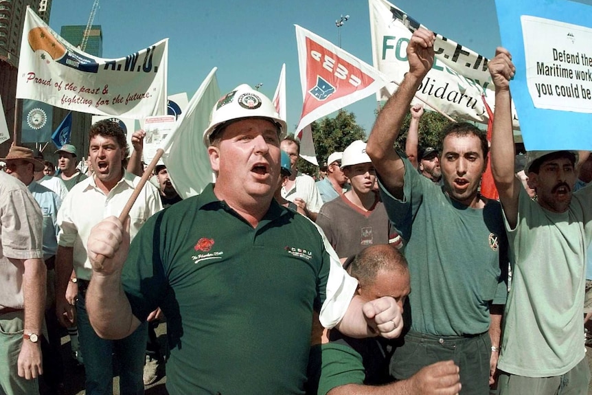 A group of men wearing hardhats and green uniforms shout and wave placards and flags.