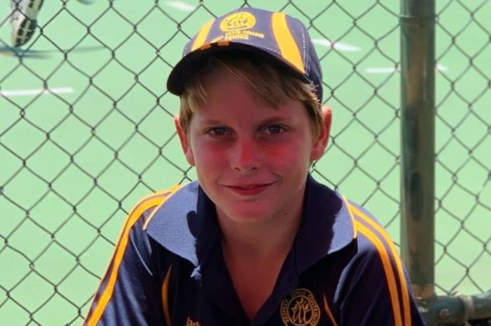 A young boy wearing a tennis uniform and cap sits in front of courts smiling.