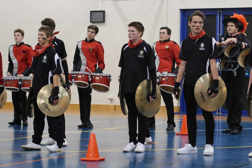 Students stand in a line holding cymbals and another row of students behind them hold three red snare drums each.