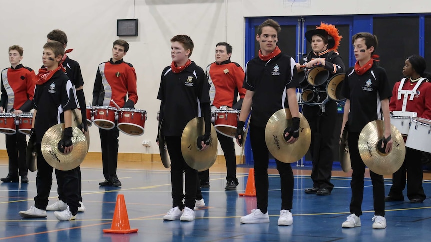 Students stand in a line holding cymbals and another row of students behind them hold three red snare drums each.