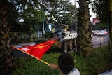 A man waves a flag next to an armoured vehicle as it drives along a civilian road past palm trees.