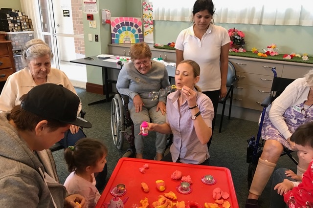 A group of elderly aged care residents watch on as a worker blows bubbles with a young child.